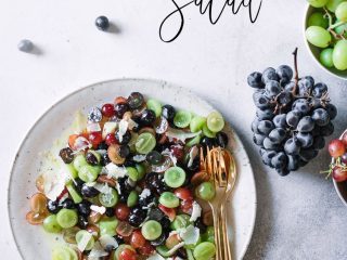 A grape fruit salad on a white plate with grapes, cheese, and wine on a white table and the words "Grape, Parm, and Pepper Fruit Salad" in black writing.