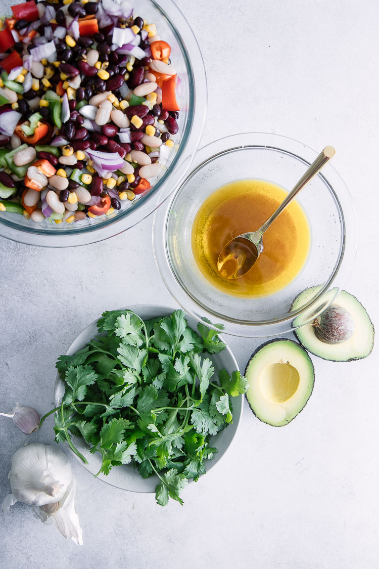 A bean salad on a white plate on a white table with olive oil, avocados, cilantro, and garlic.