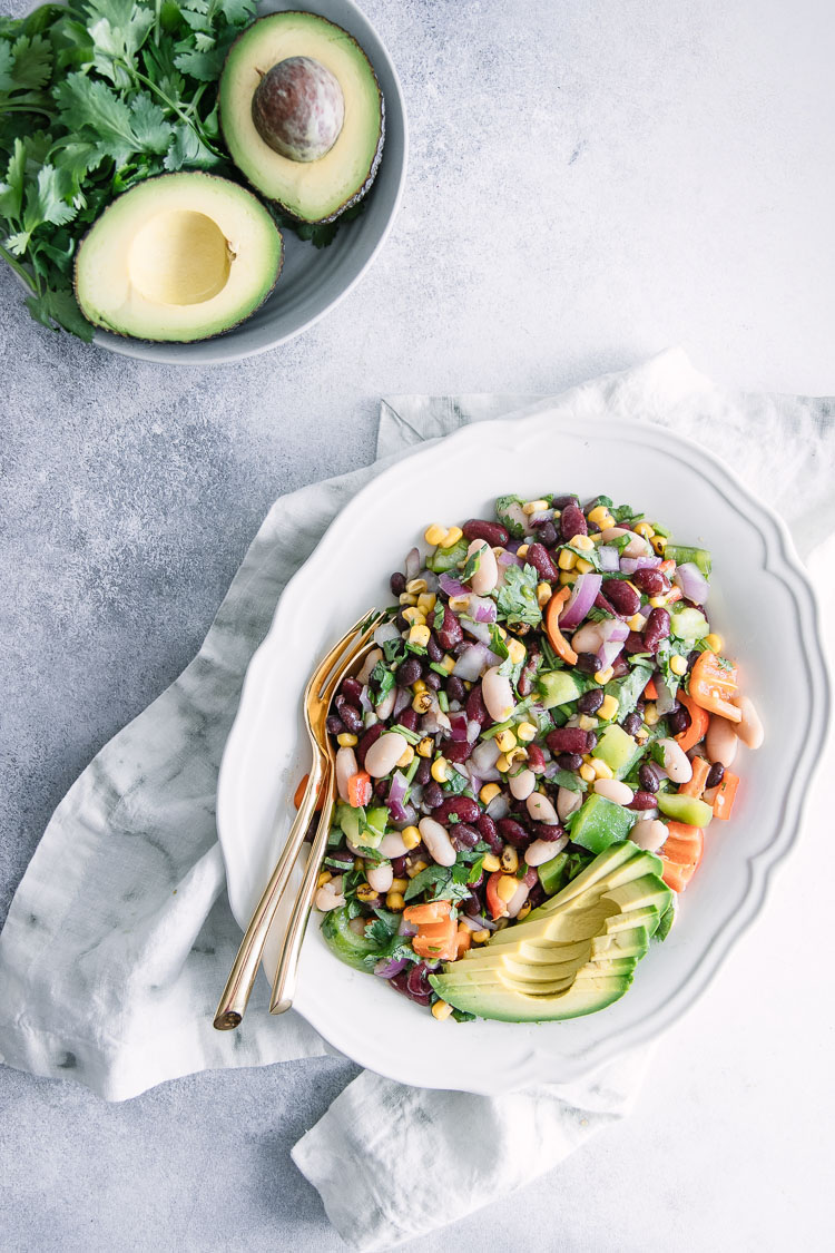 A white plate with a mixed bean salad and a gold fork on a white table with avocados and cilantro.