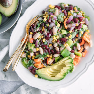 A white plate with mixed bean and vegetables salad on a blue table with a bowl of avocados.