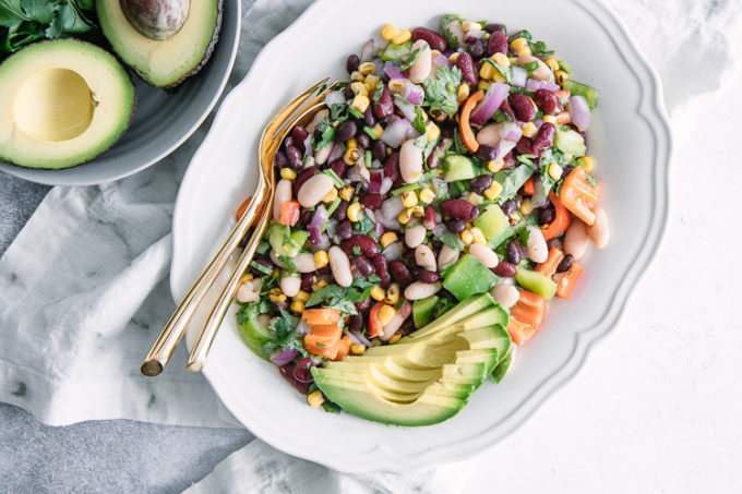 A white plate with mixed bean and vegetables salad on a blue table with a bowl of avocados.