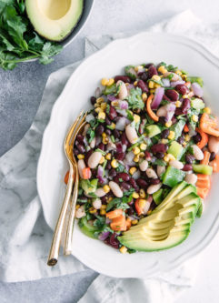 A round plate with a bean, corn, pepper, and avocado salad with a white napkin and a bowl of cilantro.