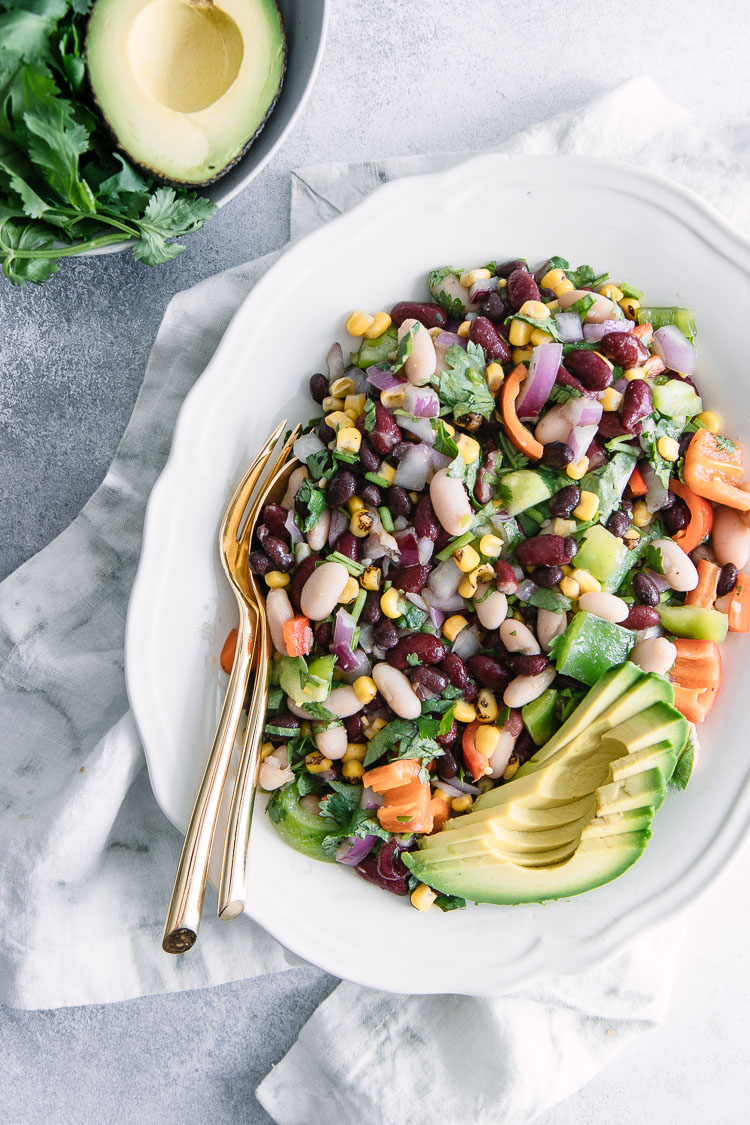 A round plate with a bean, corn, pepper, and avocado salad with a white napkin and a bowl of cilantro.