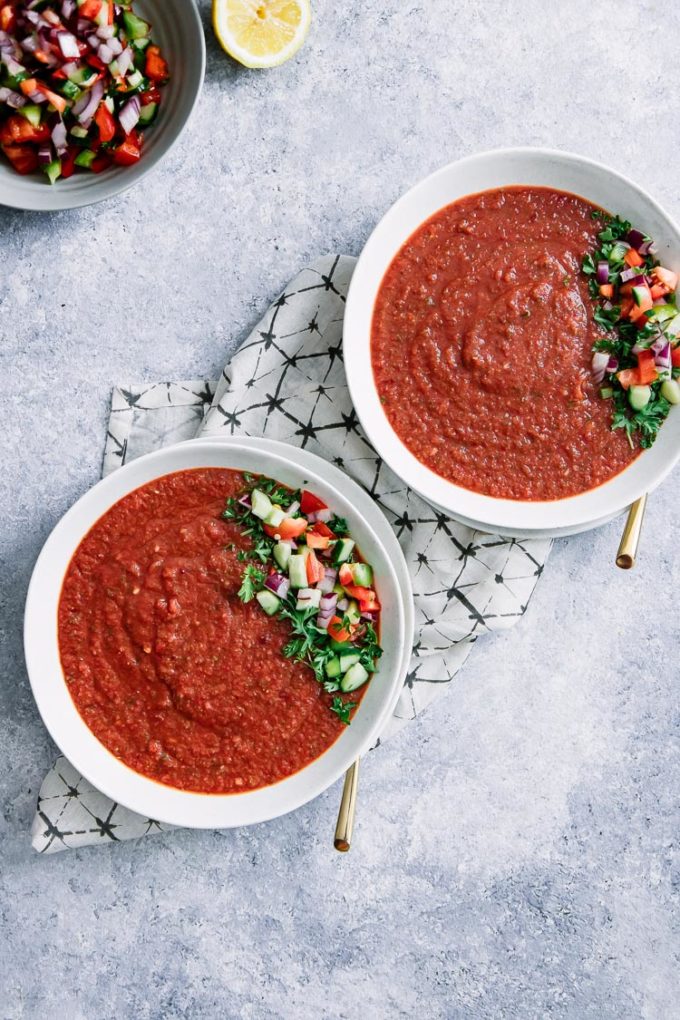 Two bowls of spicy tomato and cucumber gazpacho on a blue table with cucumber tomato salsa.