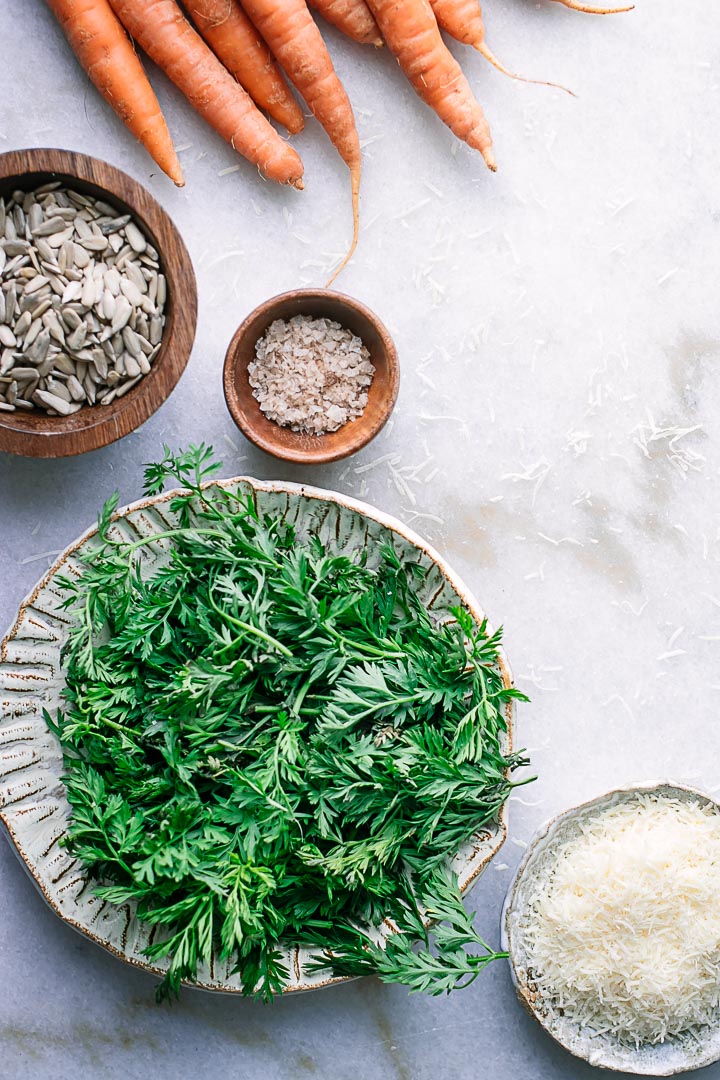 A bowl of carrot greens, a bunch of carrots, a bowl of salt, and shredded cheese on a white table.