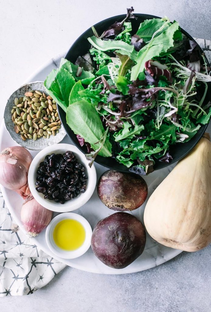 a cutting board with mixed greens, butternut squash, pumpkin seeds, shallots, beets, cranberries, and oil