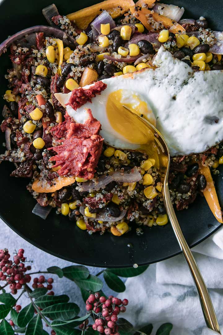 a fork breaking a sunny egg on top of a quinoa and black bean salad in a black bowl
