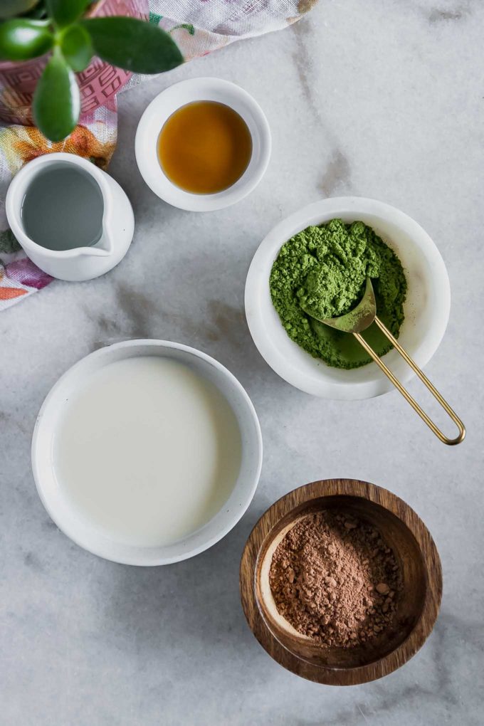 bowls of matcha, cocoa, agave nectar, water, and plant-based milk on a marble countertop