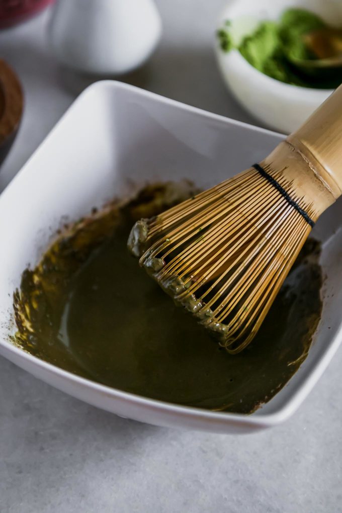 a wooden matcha whisk in a white bowl with dissolved matcha and mocha powders in water