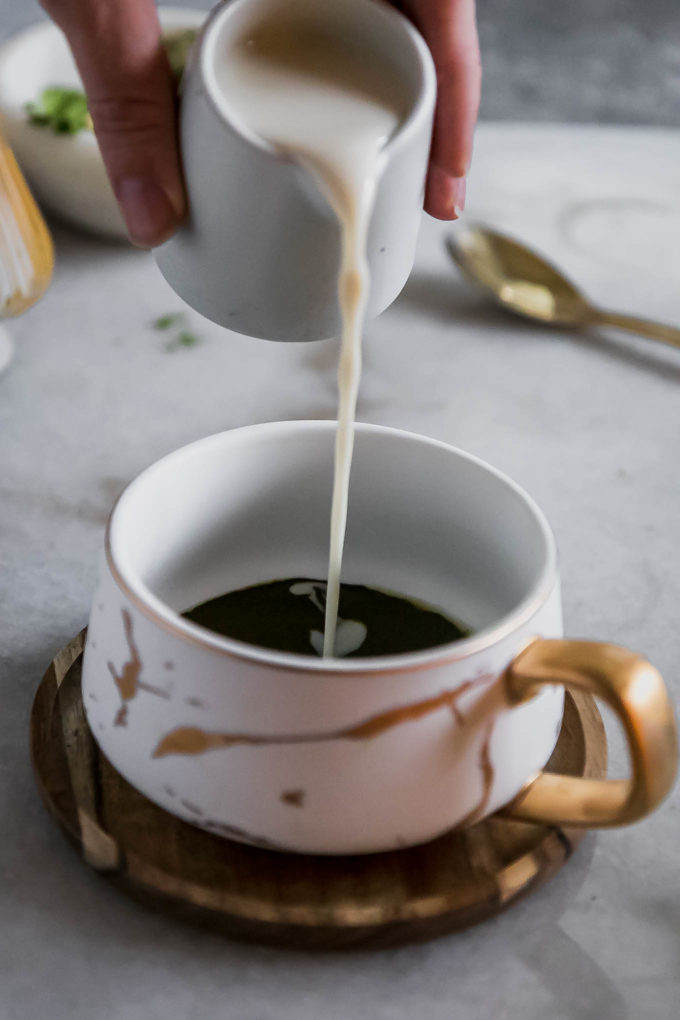 a hand pouring milk into a white mug filled with matcha tea