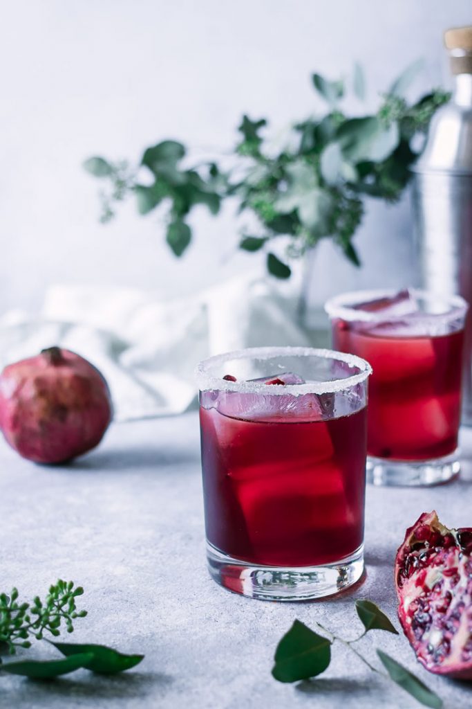 two red margaritas on a blue table with a pomegranate and green leaves