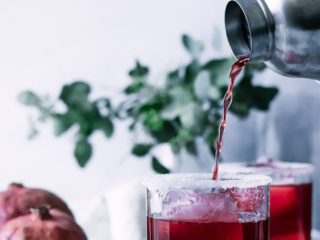 a hand pouring a red pomegranate margarita into a glass with ice and salt with pomegranates on a white table