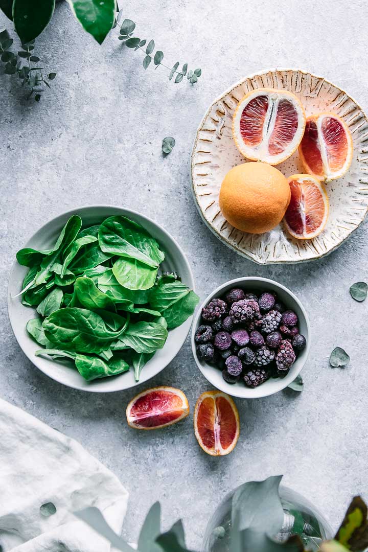 a bowl of spinach, a bowl of frozen berries, and a plate with blood oranges on a blue table