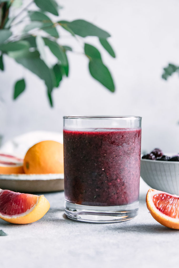 A purple smoothie in a short glass with oranges and a white napkin in the background