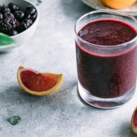 a dark purple smoothie with an orange slice on a blue table with fresh mint