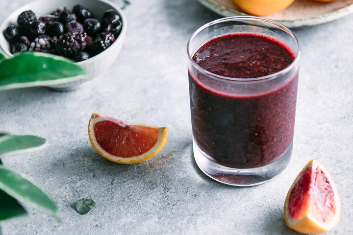 a dark purple smoothie with an orange slice on a blue table with fresh mint