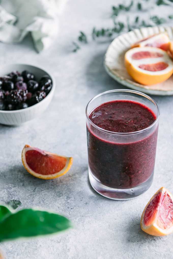 a dark red smoothie on a blue table with slices of blood orange and a bowl of blackberries and blueberries