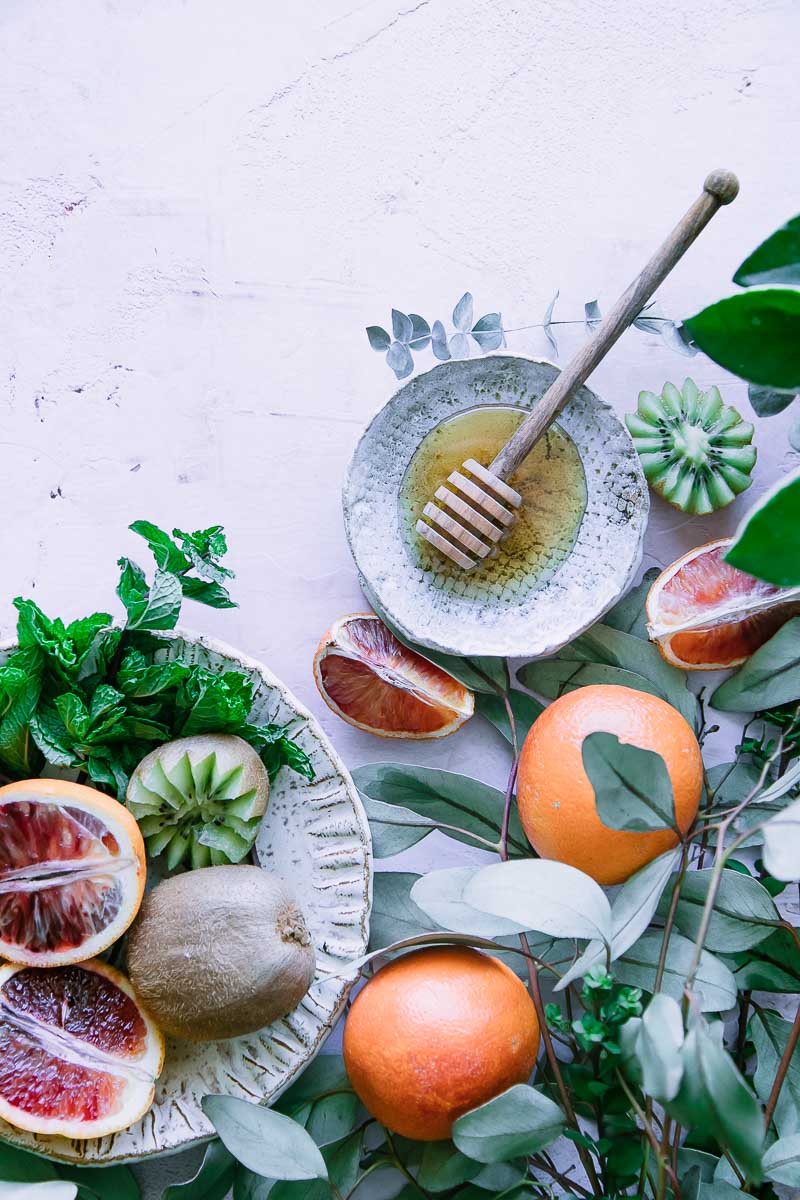 blood oranges, kiwi, and small plate of honey with a honey drizzler on a pink table with green leaves