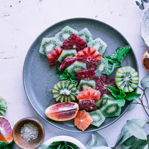 a blue plate with a fruit salad with oranges and kiwi on a table with a bowl of salt and a bowl of honey
