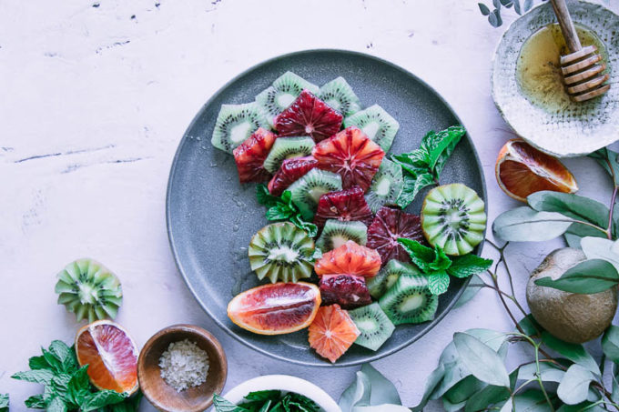 a blue plate with a fruit salad with oranges and kiwi on a table with a bowl of salt and a bowl of honey