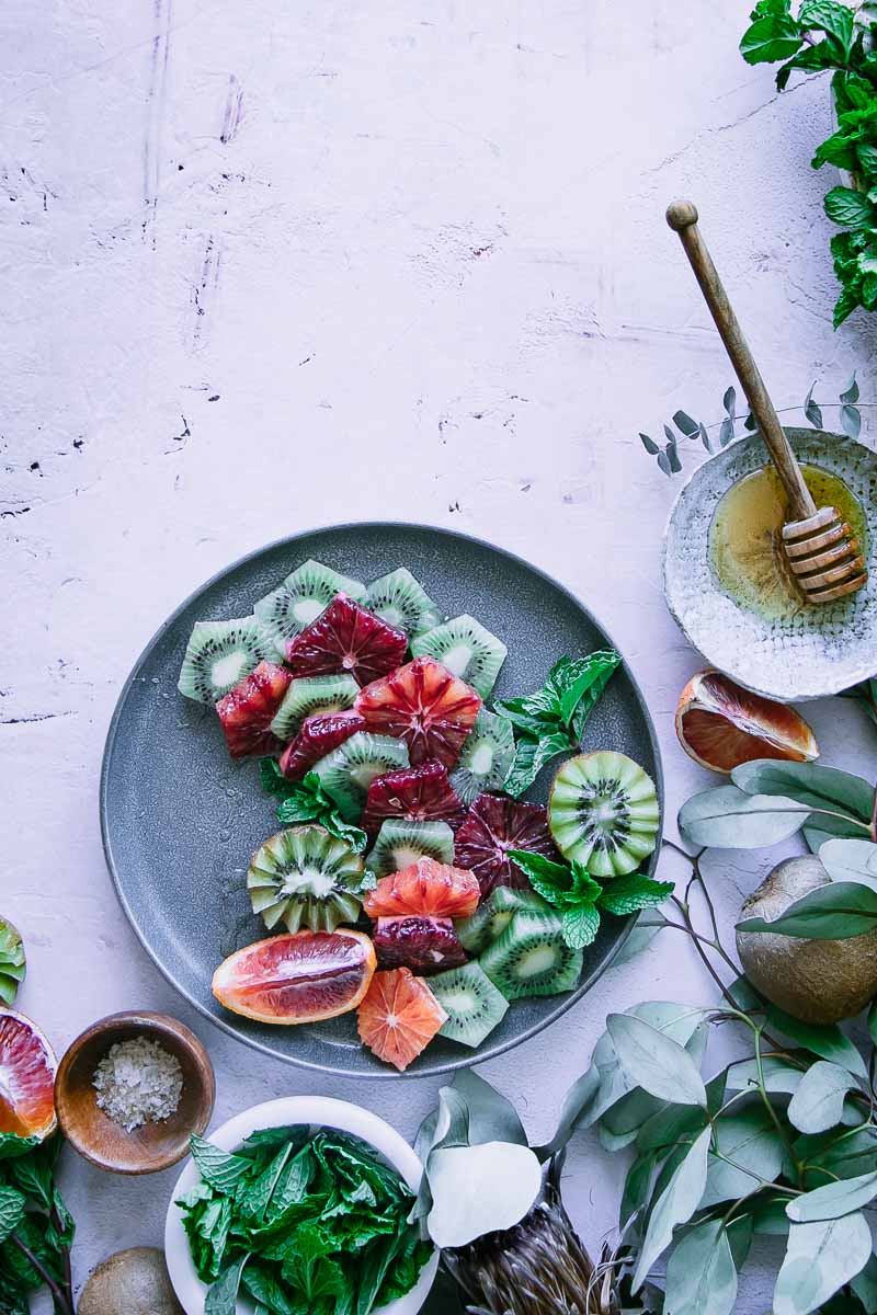 a winter citrus salad with blood oranges, kiwi, mint, honey, and sea salt on a blue plate on a pink table with honey and cut fruit