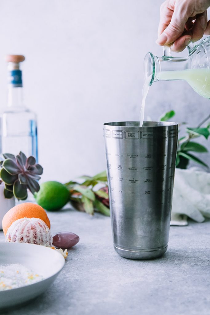 lime juice pouring into a metal cocktail shaker with a bottle of tequila in the background and oranges on a white table