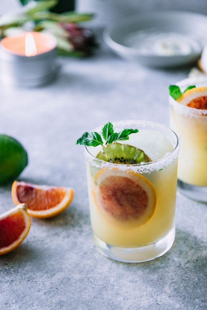 two highball glasses with margaritas, blood oranges, and kiwi and a salted rim