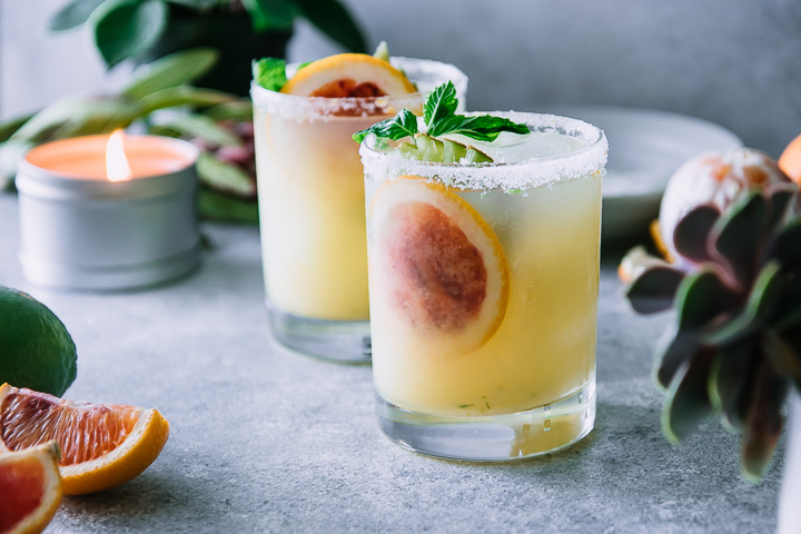 two margaritas on a blue table with sliced blood oranges and a candle with a green plant in the background