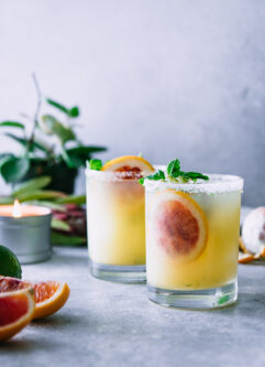two salted margaritas on a white table with plants and candles