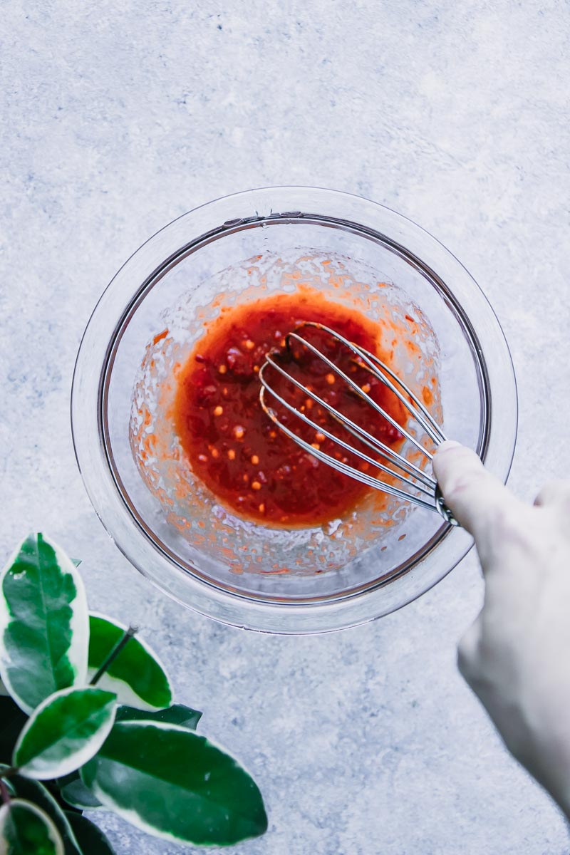 a hand stirring a red sauce in a glass mixing bowl on a blue table