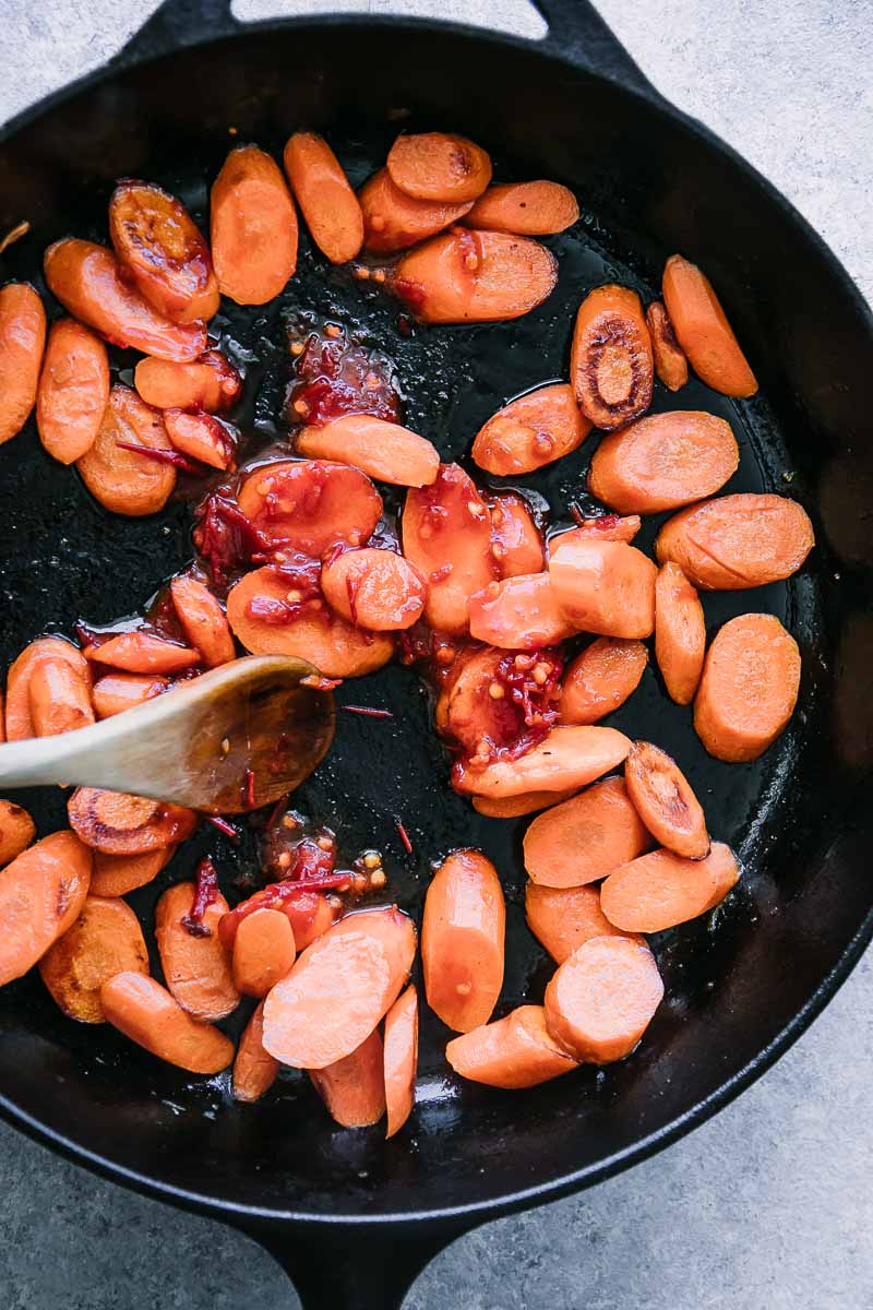 a black cast iron skillet with orange carrots and a hand stirring in a red harissa sauce