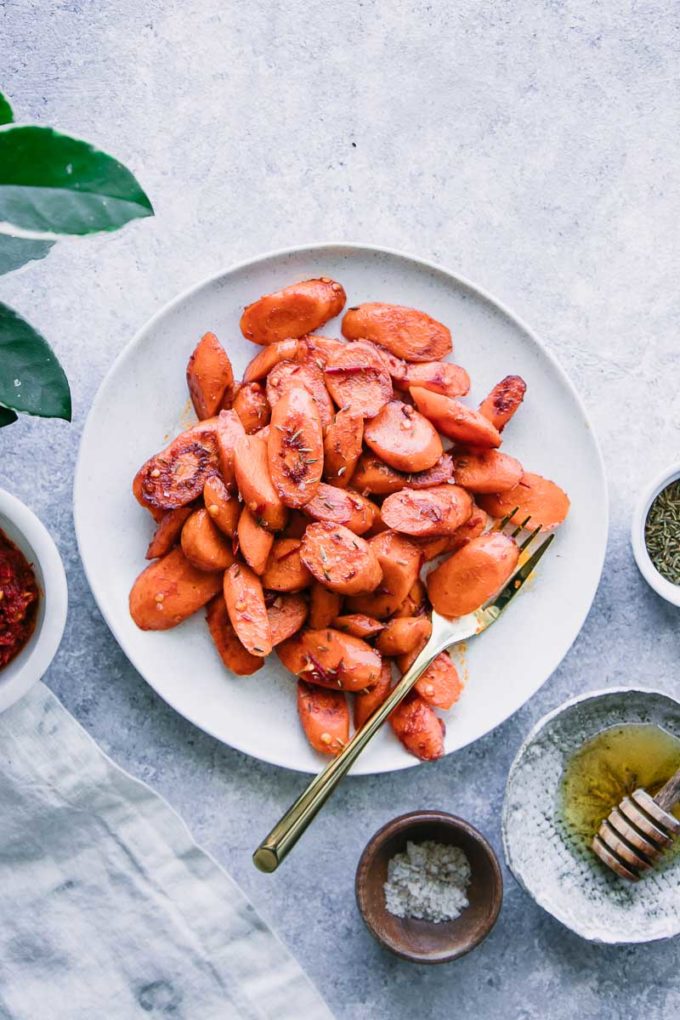 cooked carrots on a white plate with a gold fork and a side of honey and harissa sauce on a blue table