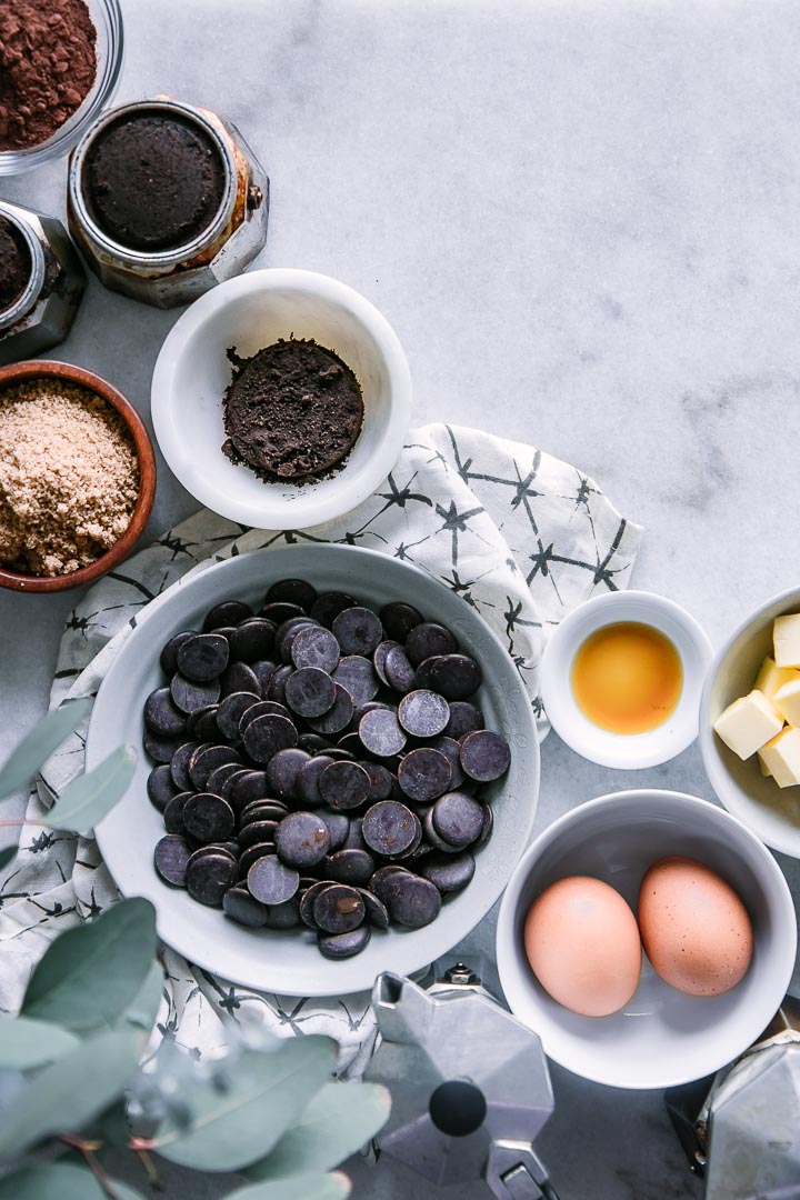 a bowl of chocolate chips, two eggs, a ramekin of vanilla, cut butter, brown sugar, and coffee grounds on a white table with a white and black napkin