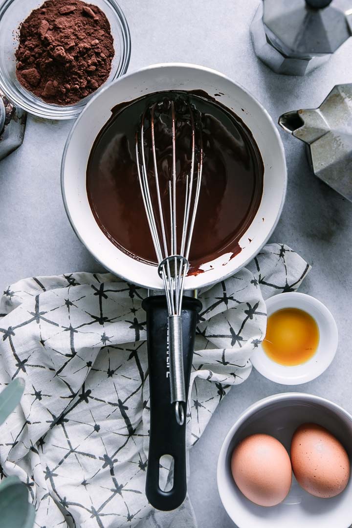 melted chocolate in a white and black pan with a wire whisk and a bowl of eggs and brown sugar on a white table
