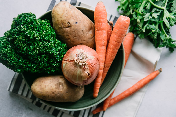 a bowl of onions, carrots, potatoes, and kale on a blue and white napkin on a white table