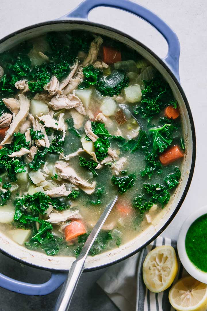 a large blue ceramic dutch oven filled with chicken kale and potato soup with a silver ladle