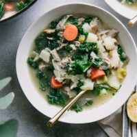 a bowl of chicken soup with potatoes, kale, and carrots on a blue table with a half a lemon