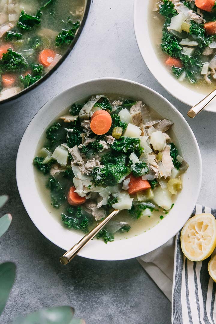 a bowl of chicken potato kale soup on a white table with flowers and a large blue soup pot in the background