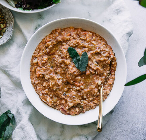 a white bowl with farro and vegetable soup on a white table with sage leaves and a white napkin