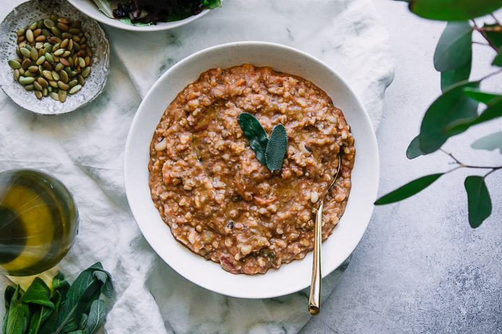 a white bowl with farro and vegetable soup on a white table with sage leaves and a white napkin