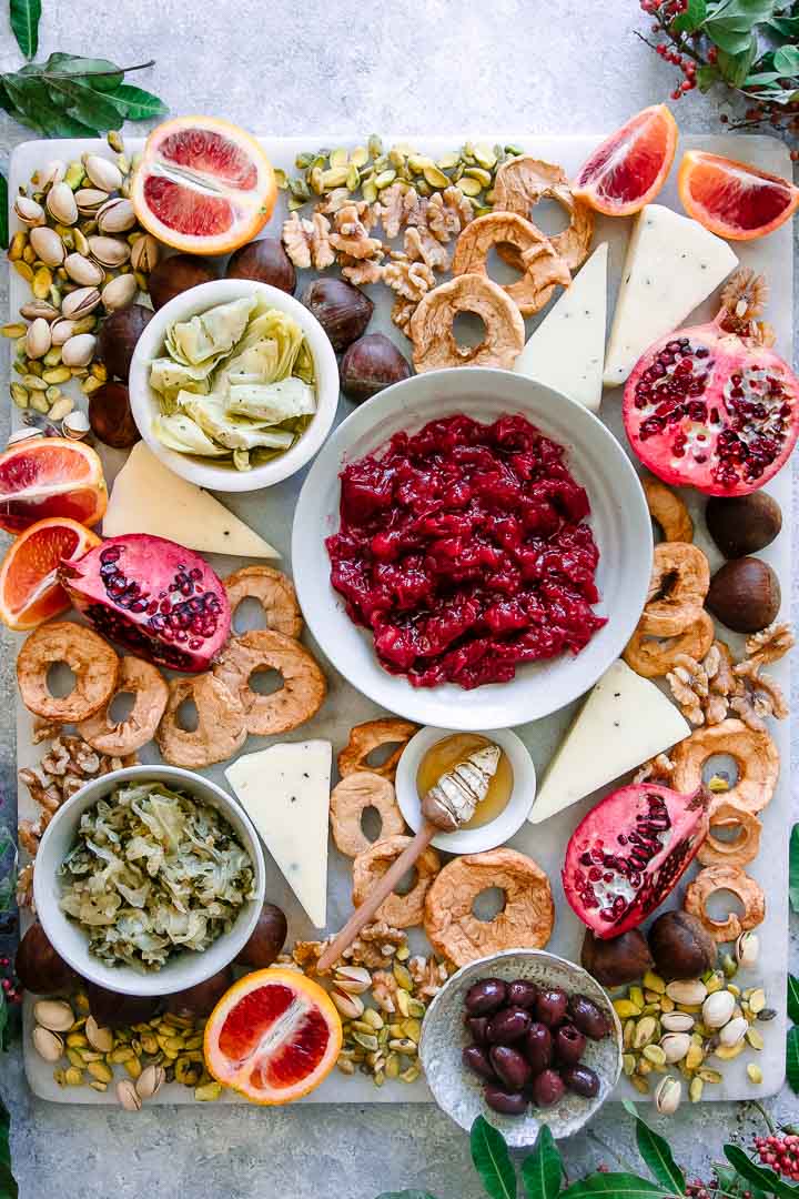 a platter with oranges, pomegranates, chestnuts, walnuts, pistachios, and olives on a white table with green leaves