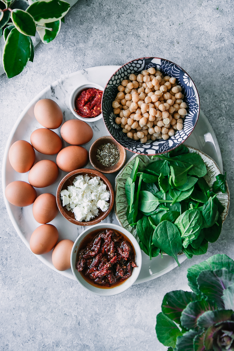 eggs, feta cheese, sun-dried tomatoes, spinach, chickpeas, sea salt, and harissa sauce on a white cutting board on a blue table