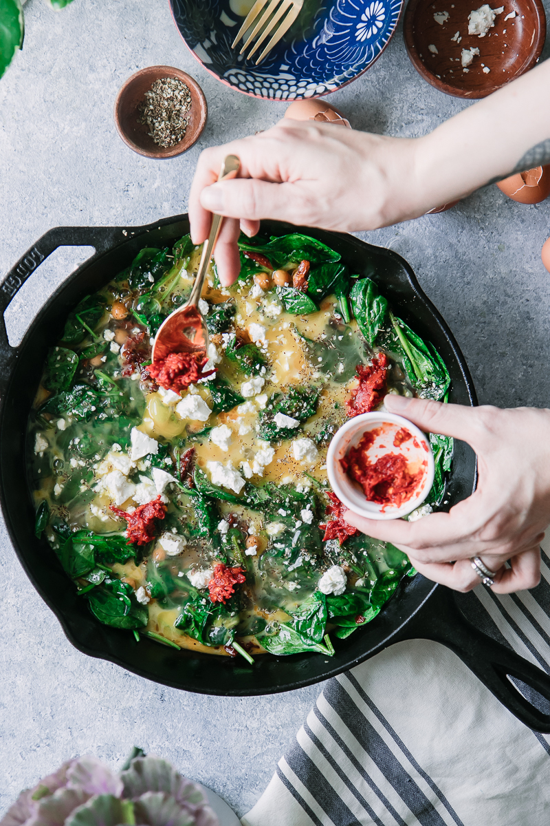 a hand spooning harissa sauce over a cast iron skillet with eggs and spinach