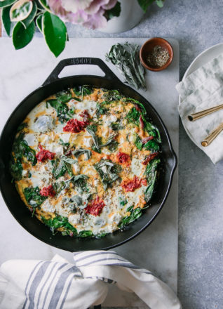 a frittata with spinach, red harrisa sauce, and feta cheese in a black cast iron skillet on a white table with a white napkin and a plate with gold forks