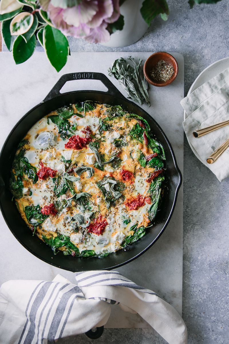 a frittata with spinach, red harrisa sauce, and feta cheese in a black cast iron skillet on a white table with a white napkin and a plate with gold forks