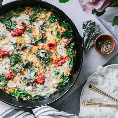 a frittata in a cast iron skillet on a white marble table with a bowl of sea salt and fresh sage leaves