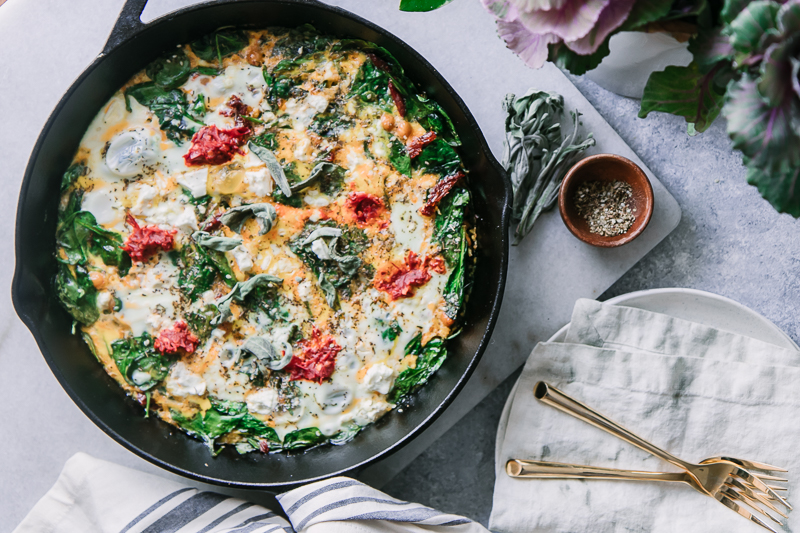 a frittata in a cast iron skillet on a white marble table with a bowl of sea salt and fresh sage leaves