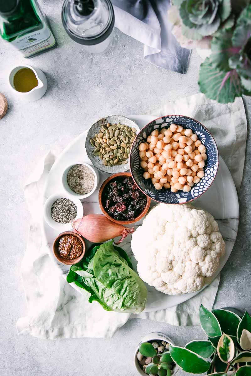 a white plate with bowls of spices, chickpeas, cranberries, and seeds on a white table with ahead of cauliflower and a bottle of olive oil