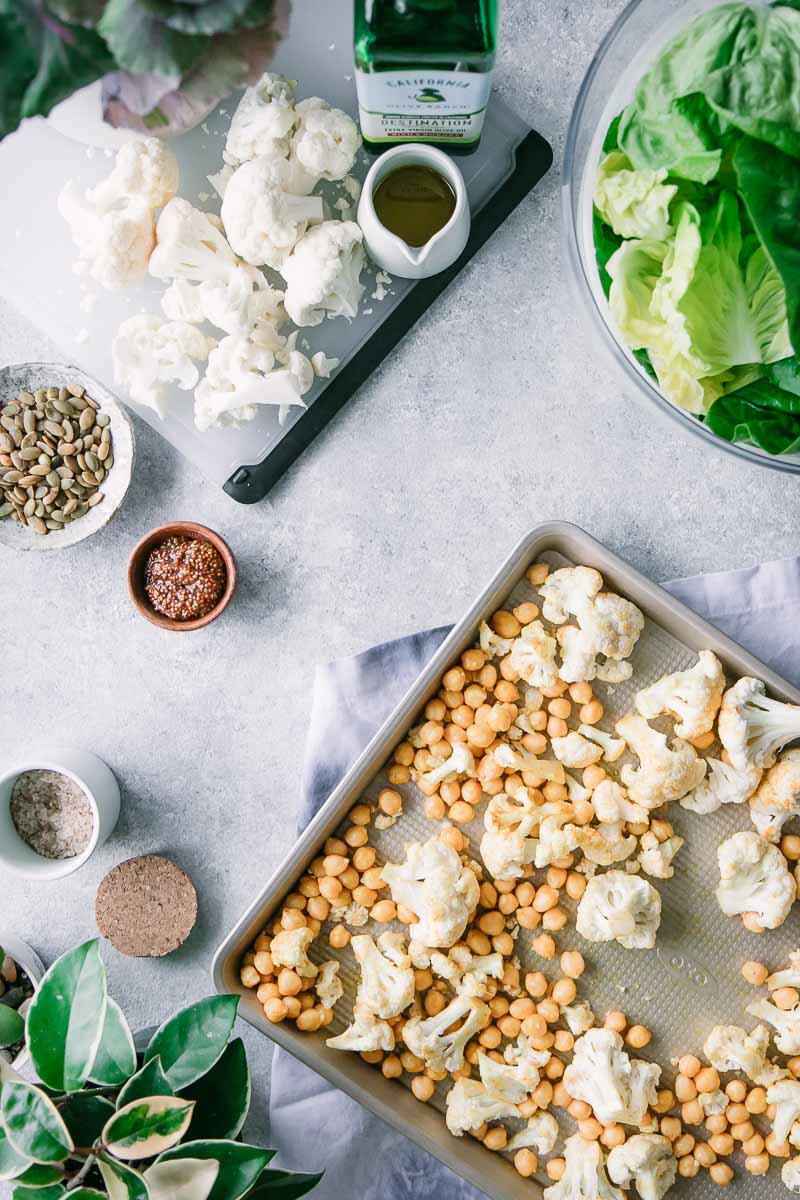 a baking sheet with roasted chickpeas and cauliflower on a white table with a cutting board and a bowl of green lettuce