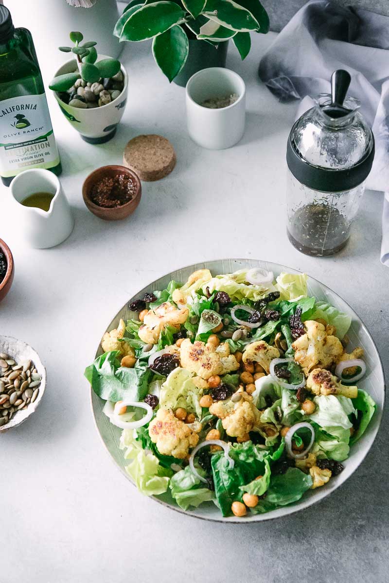 a salad with cauliflower and chickpeas on a white table with small bowls and saucers holding spices and oils